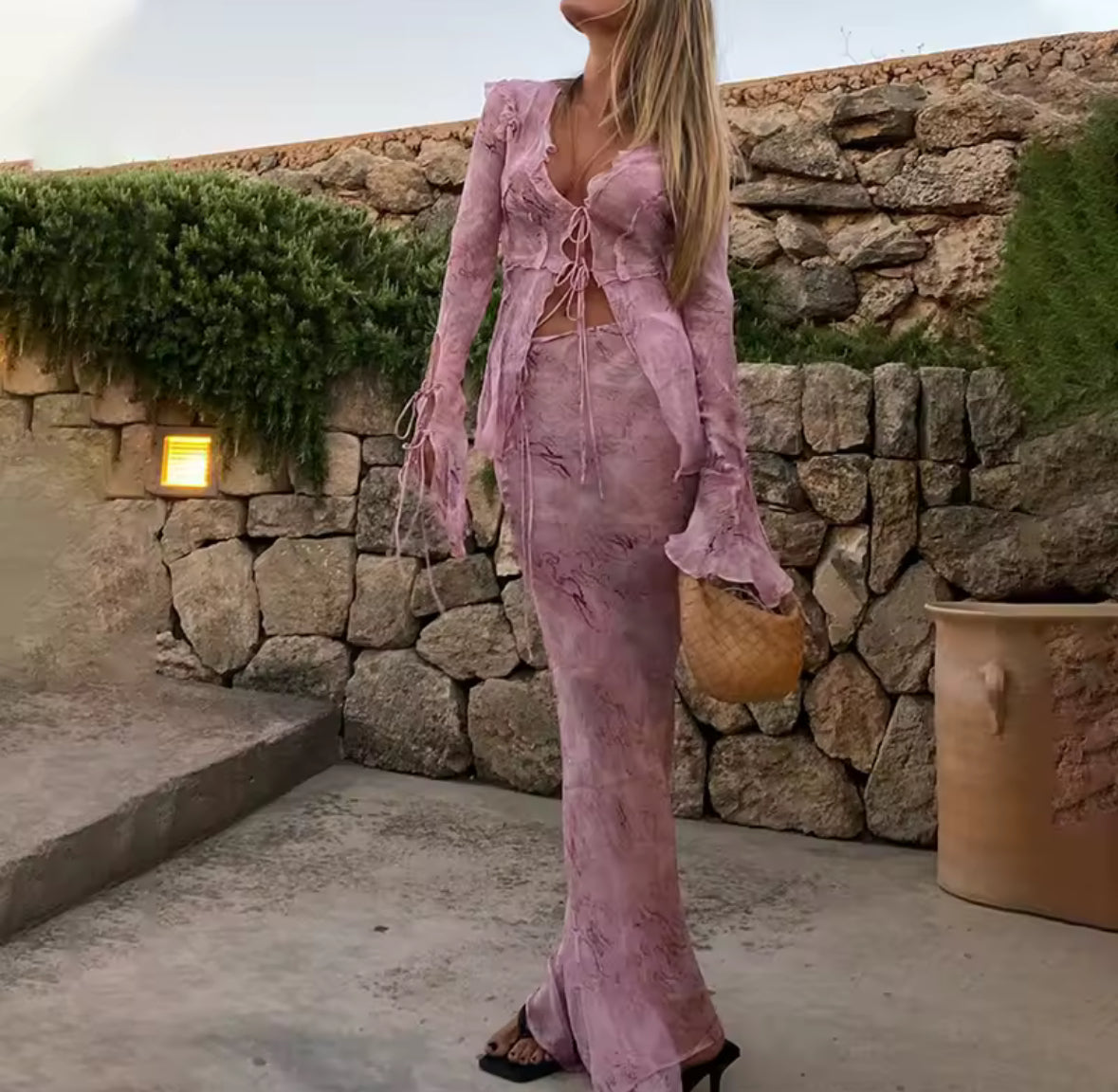 Frau trägt ein zweiteiliges fliederfarbenes Boho-Set bestehend aus Crop-Top mit Schnürung und Maxi-Rock vor einer Steinmauer, kombiniert mit geflochtener Tasche und High Heels – sommerlicher Urlaubslook im Ibiza-Stil.