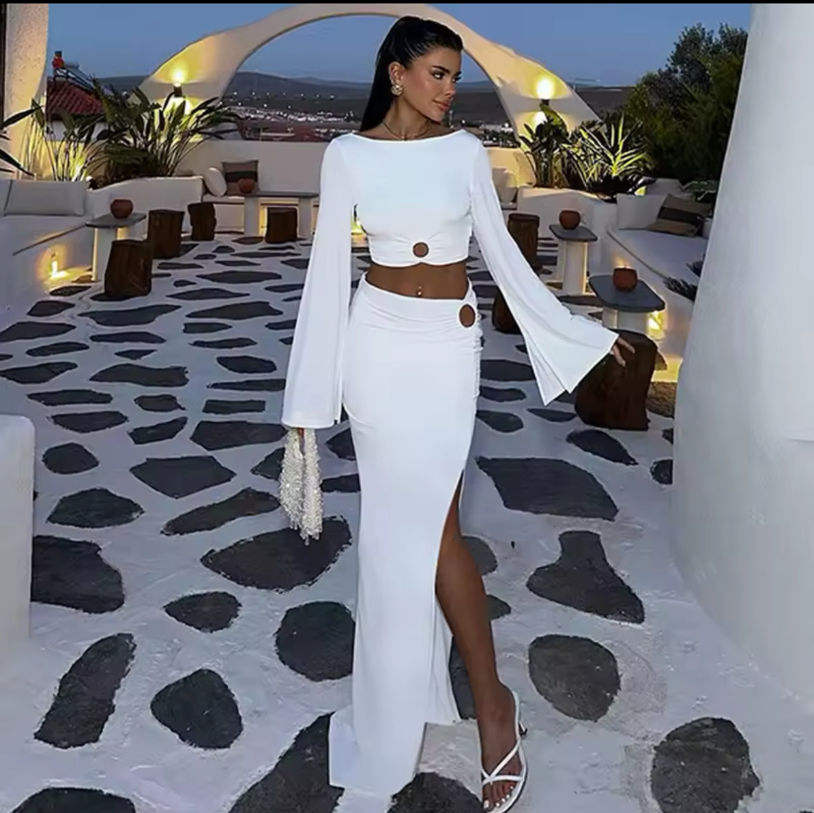 Frau in einem weißen, figurbetonten Set mit goldenen Cut-Out-Details, stehend auf einer mediterranen Terrasse