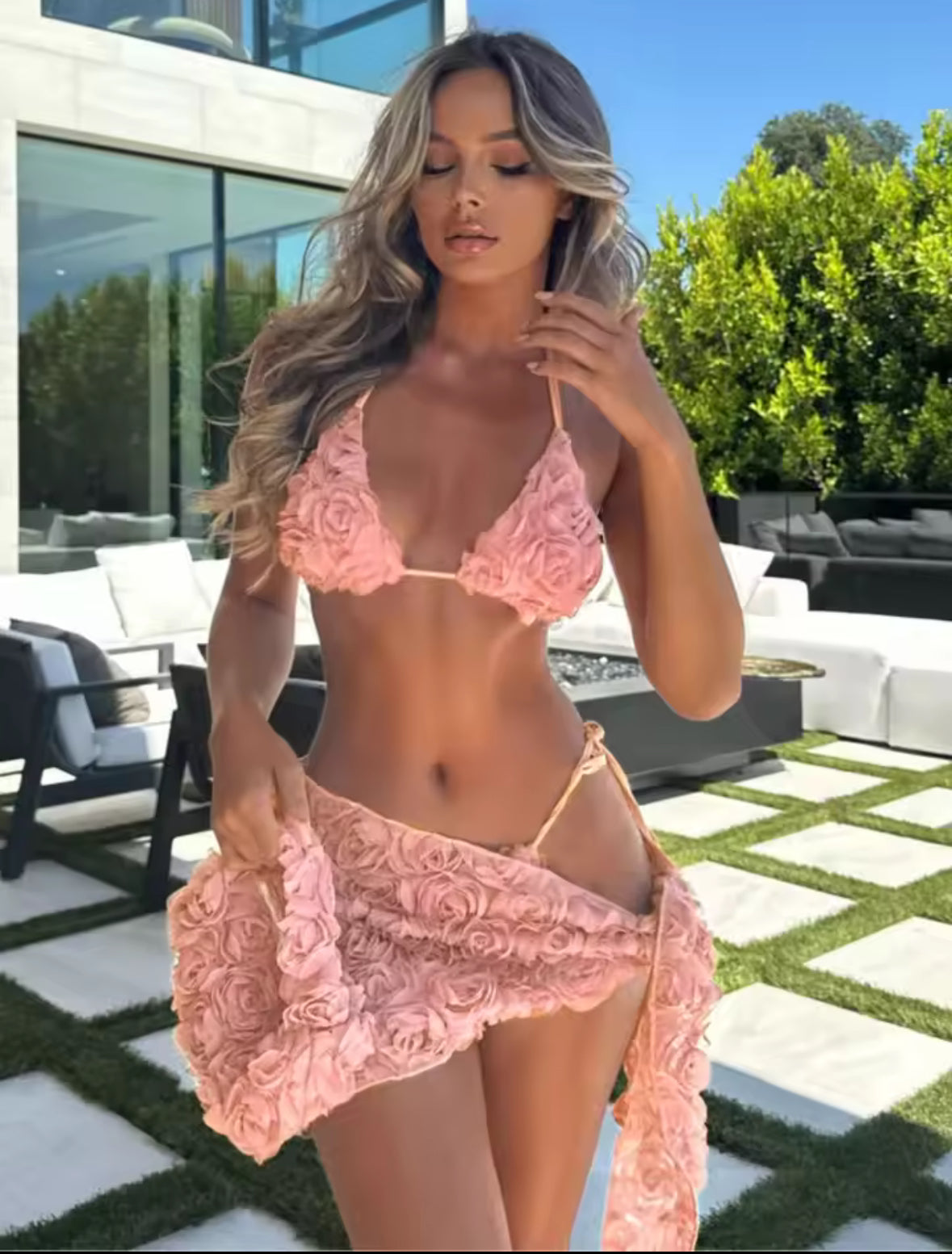Roséfarbenes Bikini-Set mit 3D-Rosenstruktur auf einem cremefarbenen Sessel unter freiem Himmel. Das Set besteht aus Triangel-Top und passendem Sarong und wird in natürlichem Licht vor einem ruhigen Poolbereich präsentiert.