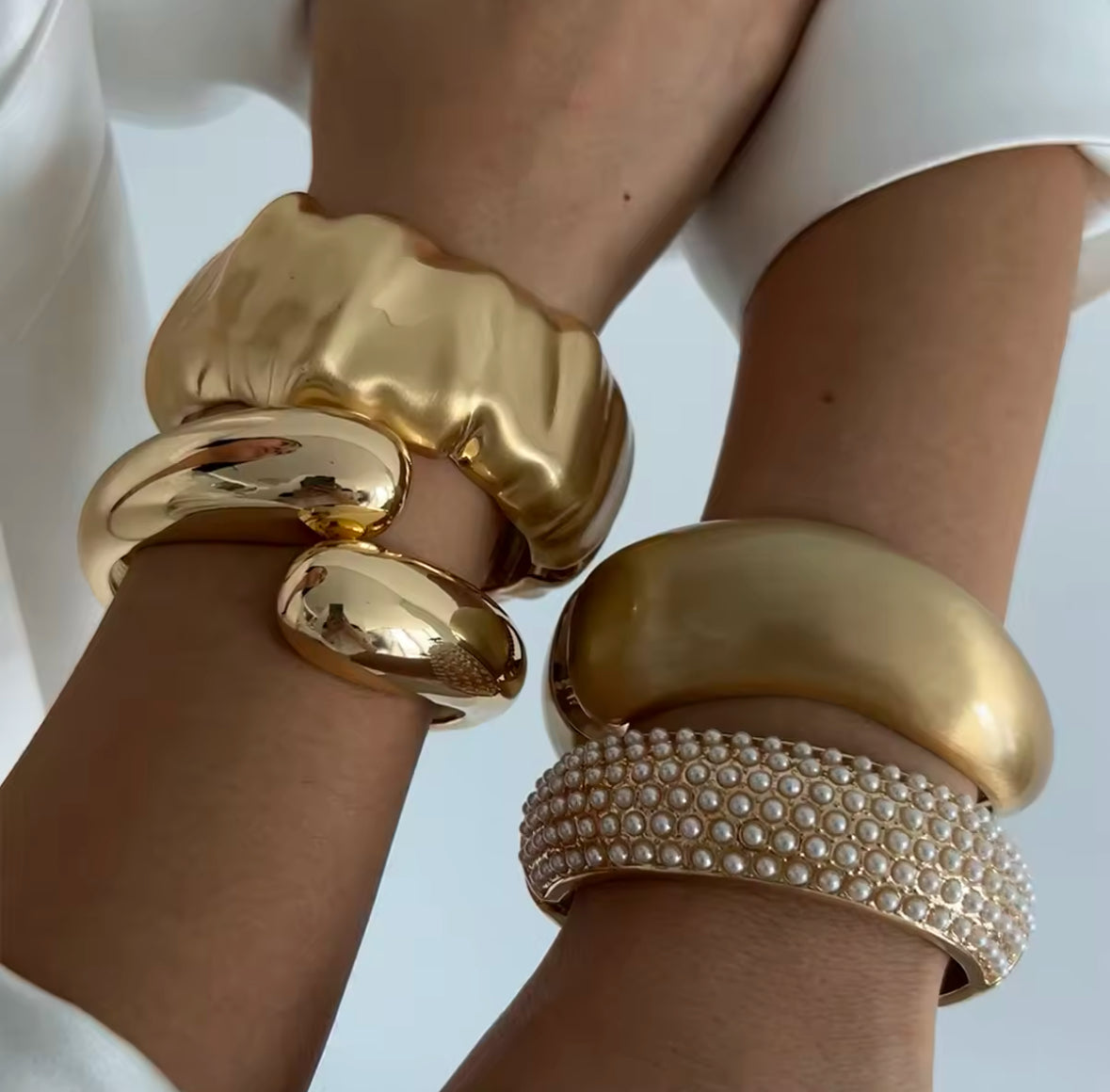 Exklusive goldene Armreifen-Kombination mit skulpturalem Design, darunter ein strukturiertes Statement-Armband, ein minimalistischer Goldreif und ein luxuriöses Perlenarmband. Hochwertige Schmuckstücke für moderne Eleganz und stilvolle Outfits.