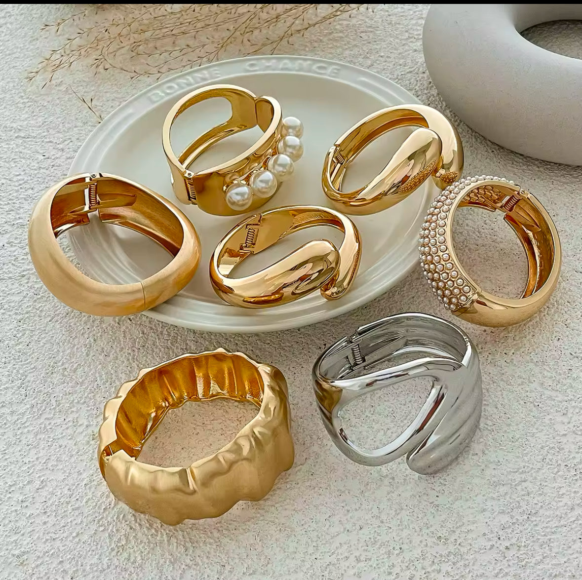 Exklusive Kollektion luxuriöser Statement-Armreifen in Gold und Silber, präsentiert auf einem eleganten Keramikteller. Verschiedene Designs mit Perlen, strukturierten Oberflächen und organischen Formen für einen modernen, stilvollen Look. Perfekt für elegante Outfits, besondere Anlässe und zeitlose Schmuck-Kombinationen.