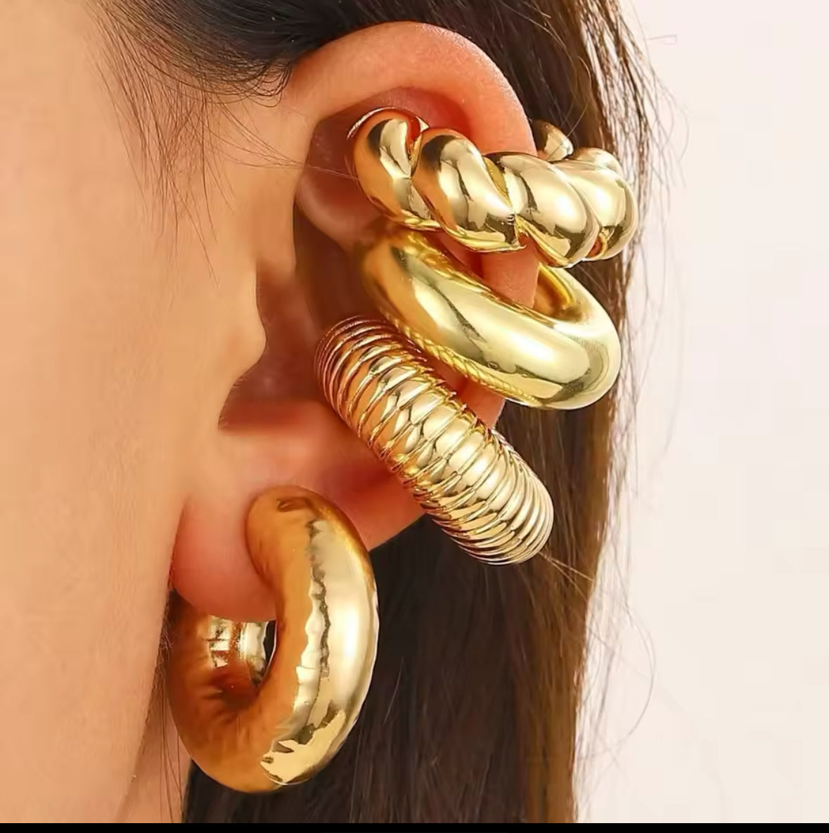 Nahaufnahme eines luxuriösen Ear-Cuff-Ohrrings in glänzendem Gold. Das mehrteilige Design umfasst verschiedene Creolen mit glatten, geflochtenen und geriffelten Oberflächen, die sich elegant um das Ohr legen. Ein auffälliges, stilvolles Statement-Piece mit modernem Touch.