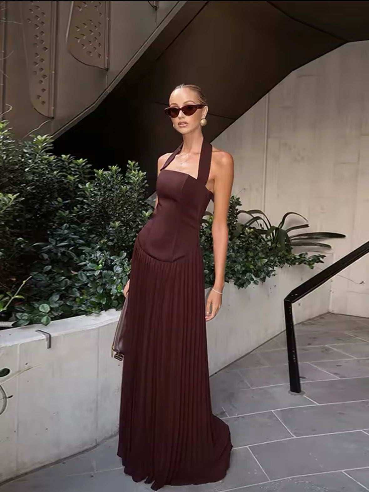 Frau trägt ein elegantes, rückenfreies Abendkleid in Espresso-Braun mit Halterneck-Trägern, figurbetontem Oberteil im Corsagenstil und fließendem Plisseerock – perfekter Look für Events, Dinners oder festliche Anlässe.