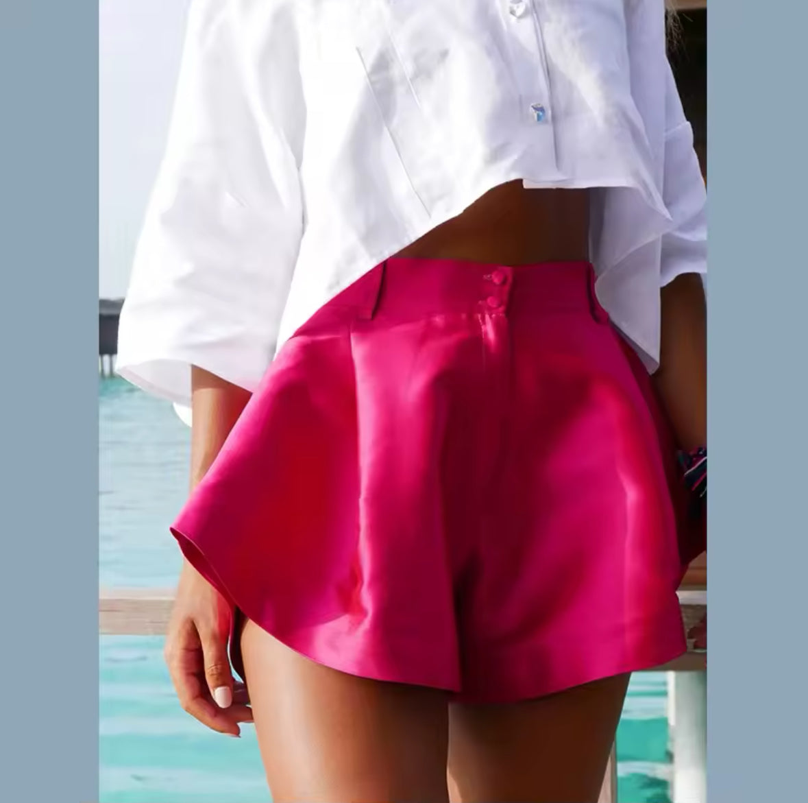 Fuchsiafarbene Satin-Shorts mit seidigem Glanz, kombiniert mit einer weißen Crop-Bluse – der perfekte Look für einen stilvollen Sommerauftritt.
