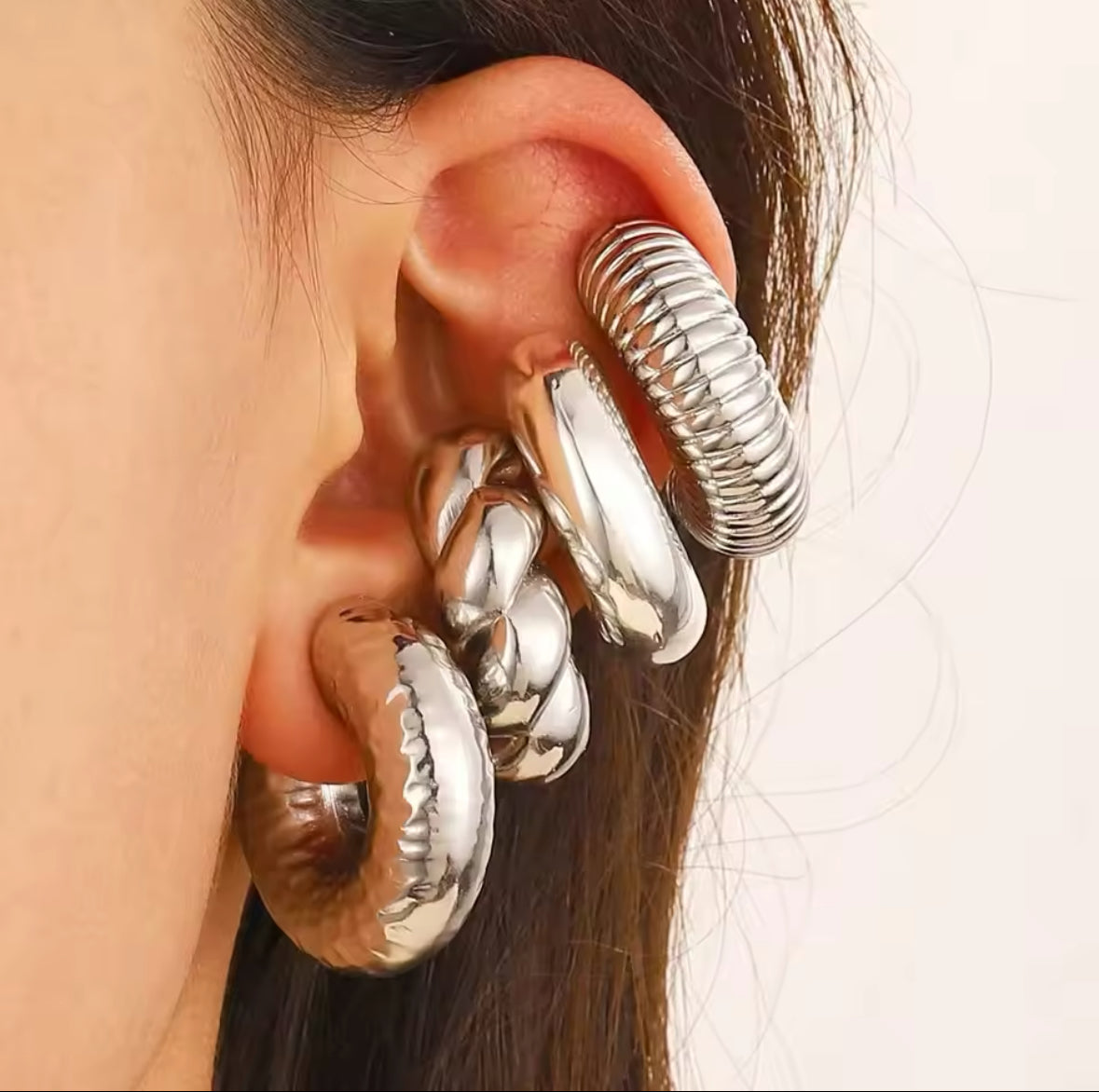 Nahaufnahme eines auffälligen Ear-Cuff-Ohrrings in glänzendem Silber. Das Set besteht aus mehreren markanten Creolen und strukturierten Elementen, die das gesamte Ohr umschließen. Das Design kombiniert glatte, gedrehte und geriffelte Strukturen für einen modernen, avantgardistischen Look.