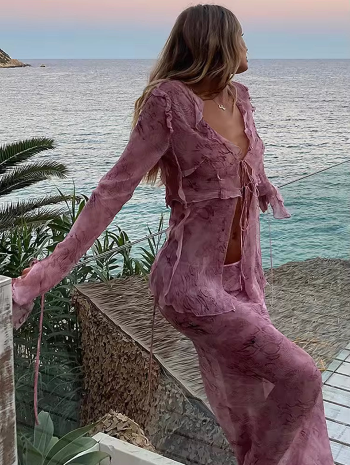 Frau trägt ein zweiteiliges fliederfarbenes Boho-Set bestehend aus Crop-Top mit Schnürung und Maxi-Rock vor einer Steinmauer, kombiniert mit geflochtener Tasche und High Heels – sommerlicher Urlaubslook im Ibiza-Stil.