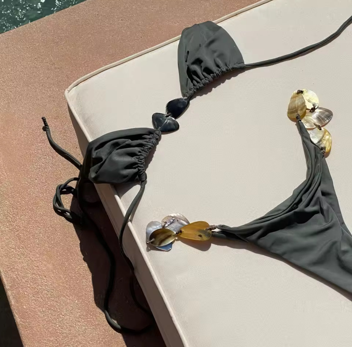 Frau trägt ein dreiteiliges Bikini-Set in Meeresfarben mit Muscheldetails und transparentem Sarong vor Steinwand.