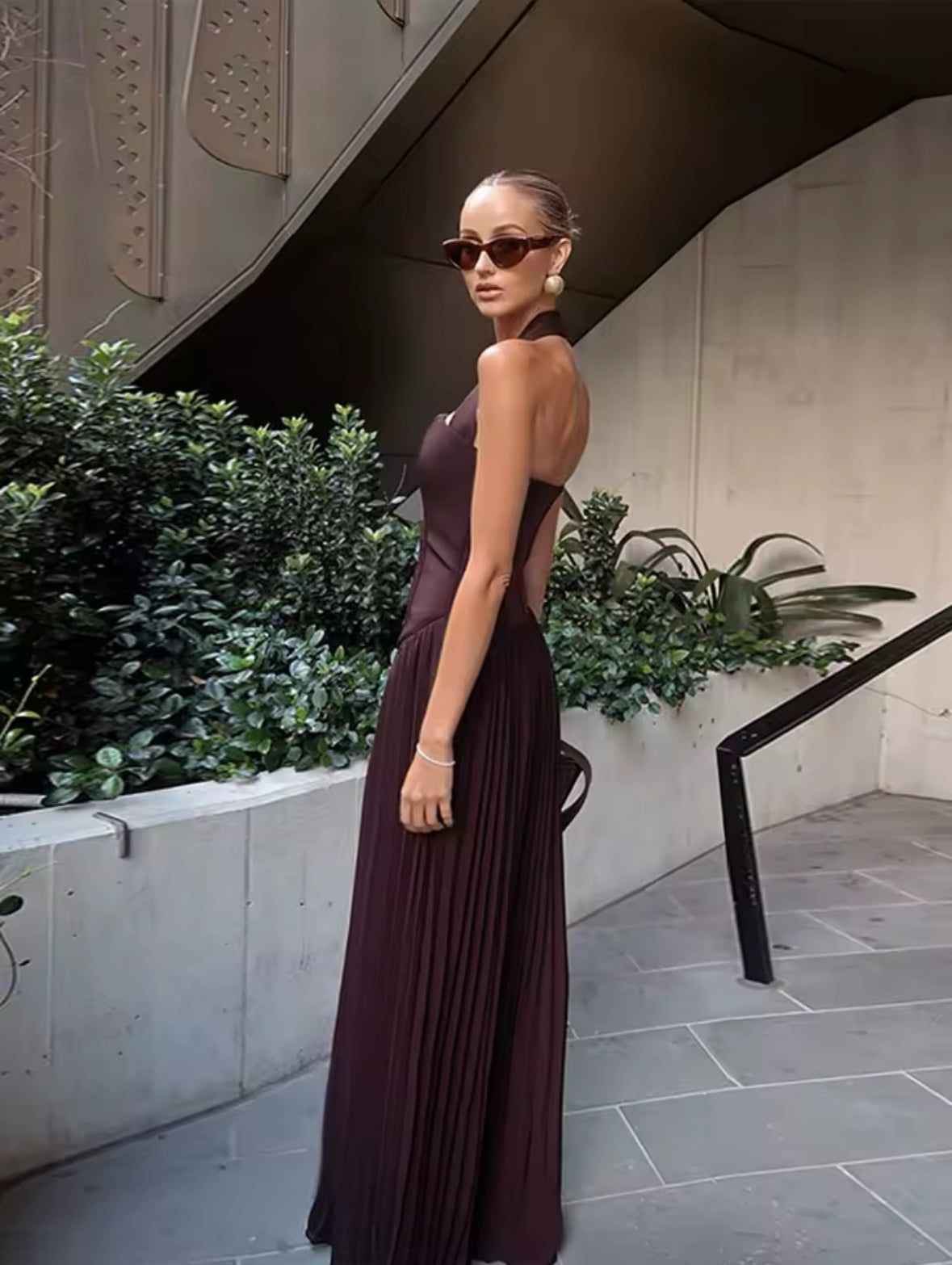 Frau trägt ein elegantes, rückenfreies Abendkleid in Espresso-Braun mit Halterneck-Trägern, figurbetontem Oberteil im Corsagenstil und fließendem Plisseerock – perfekter Look für Events, Dinners oder festliche Anlässe.