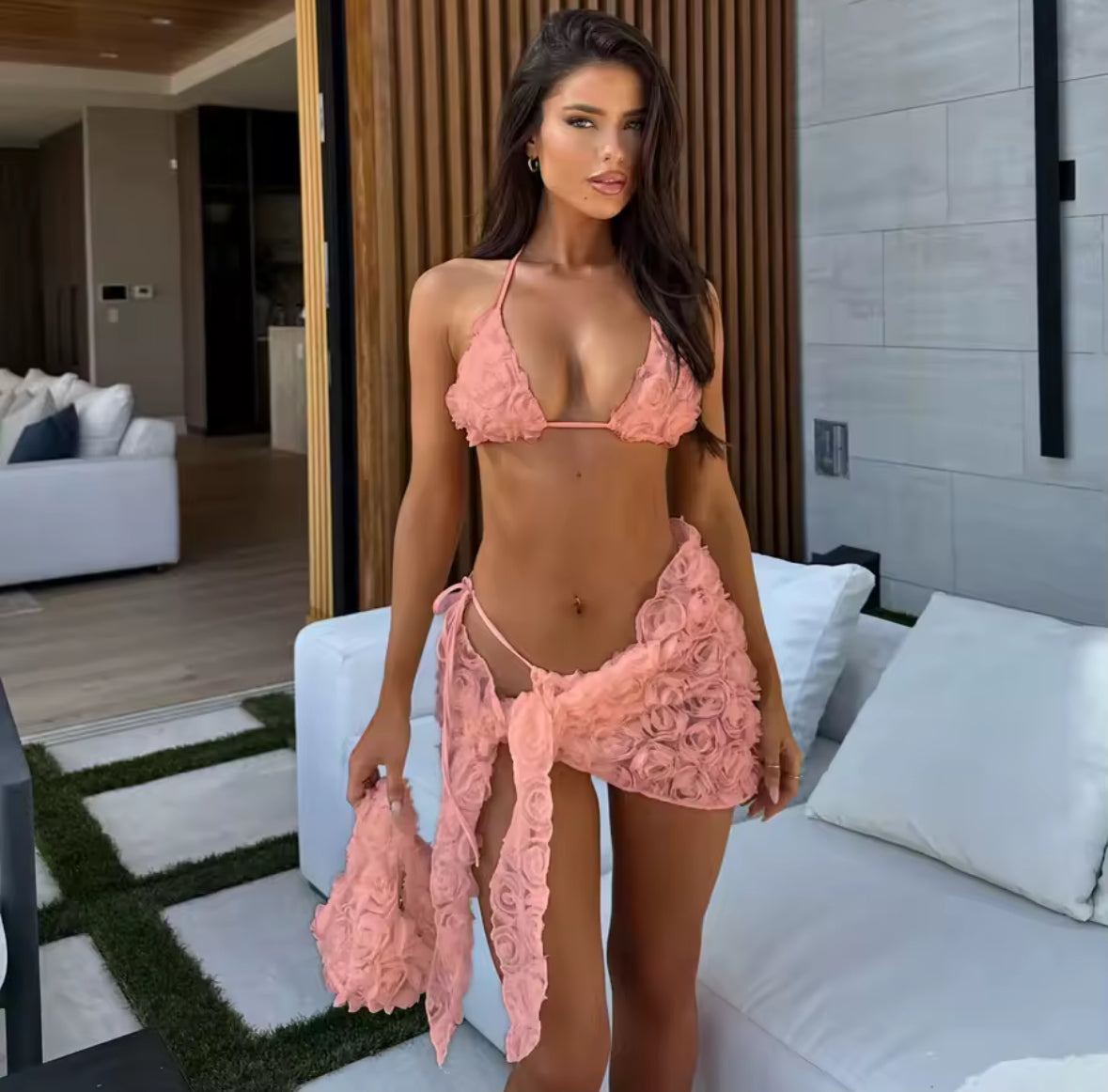 Roséfarbenes Bikini-Set mit 3D-Rosenstruktur auf einem cremefarbenen Sessel unter freiem Himmel. Das Set besteht aus Triangel-Top und passendem Sarong und wird in natürlichem Licht vor einem ruhigen Poolbereich präsentiert.