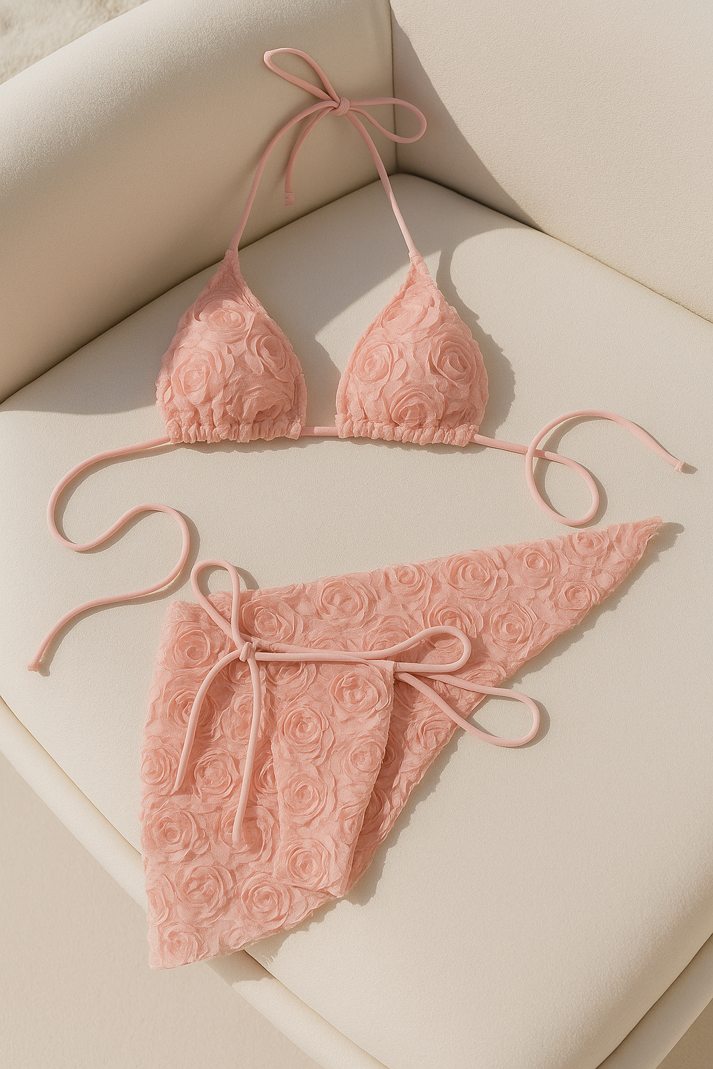 Roséfarbenes Bikini-Set mit 3D-Rosenstruktur auf einem cremefarbenen Sessel unter freiem Himmel. Das Set besteht aus Triangel-Top und passendem Sarong und wird in natürlichem Licht vor einem ruhigen Poolbereich präsentiert.