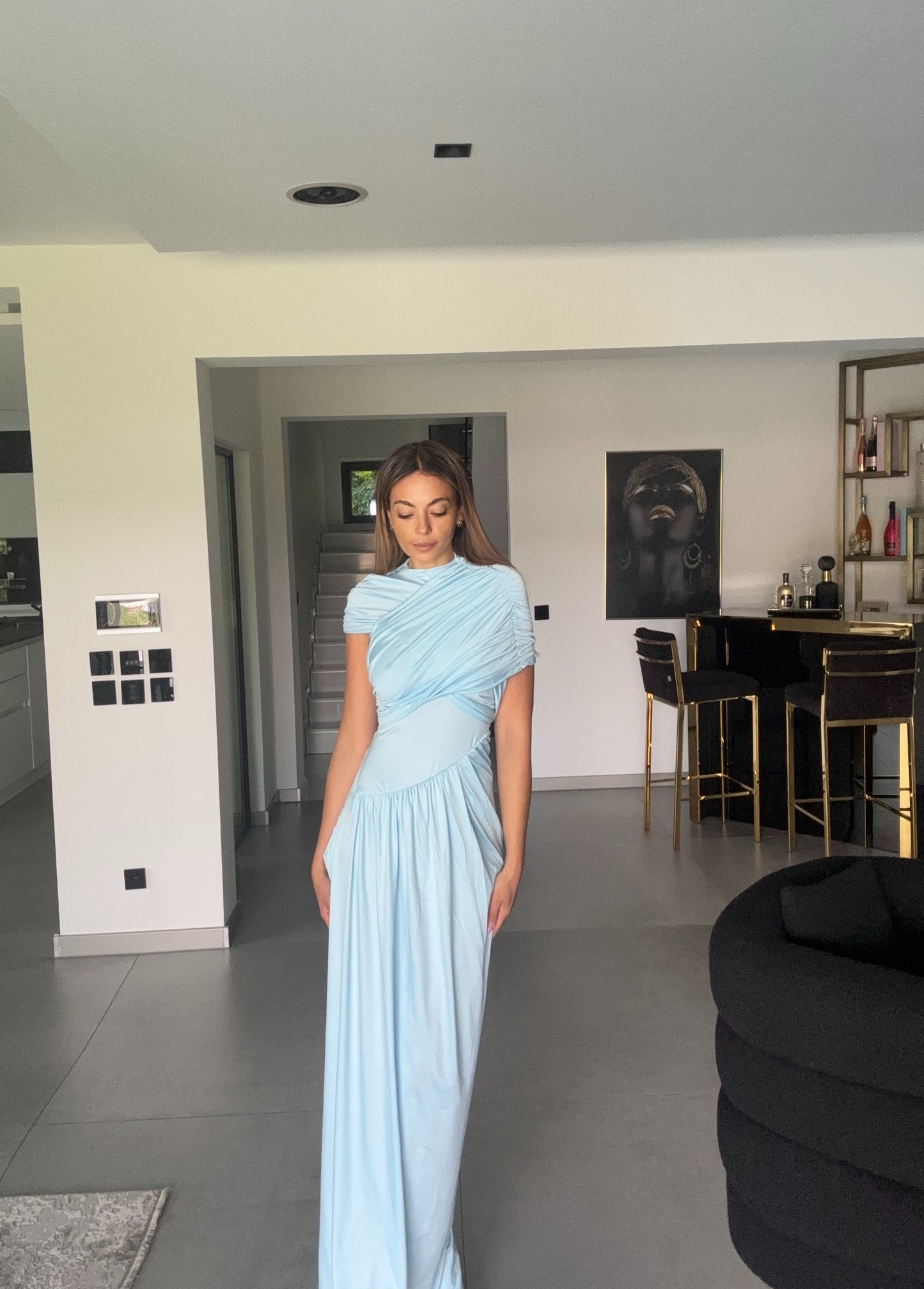 Frau trägt bodenlanges hellblaues Kleid mit drapiertem Oberteil und asymmetrischem Schnitt, steht in elegantem Interieur