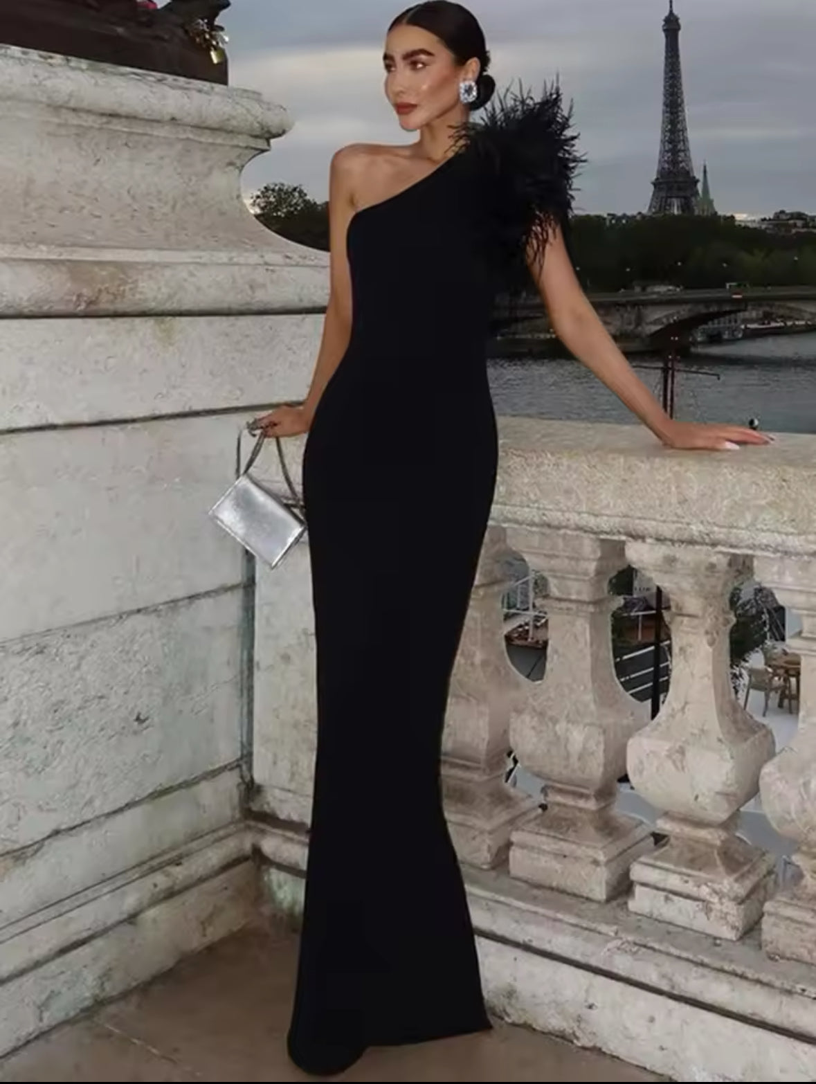 Schwarzes, bodenlanges Abendkleid mit asymmetrischem One-Shoulder-Schnitt und schwarzem Federbesatz an der Schulter – getragen vor Pariser Kulisse mit Eiffelturm im Hintergrund.
