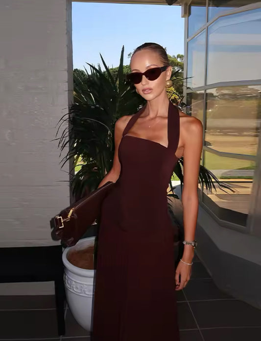 Frau trägt ein elegantes, rückenfreies Abendkleid in Espresso-Braun mit Halterneck-Trägern, figurbetontem Oberteil im Corsagenstil und fließendem Plisseerock – perfekter Look für Events, Dinners oder festliche Anlässe.
