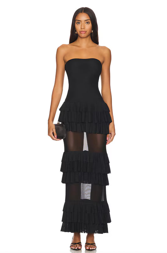Model trägt ein trägerloses schwarzes Maxikleid mit mehreren Lagen Volants und transparenten Mesh-Einsätzen im Rockbereich. Das Kleid betont die Taille und wirkt elegant und feminin.