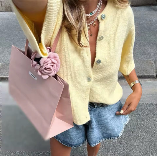 Frau trägt einen gelben Strick-Cardigan mit halblangen Ärmeln, kombiniert mit Jeans-Shorts, mehreren Ketten und einer rosa Einkaufstasche mit Stoffblume – sommerlicher Streetstyle-Look.