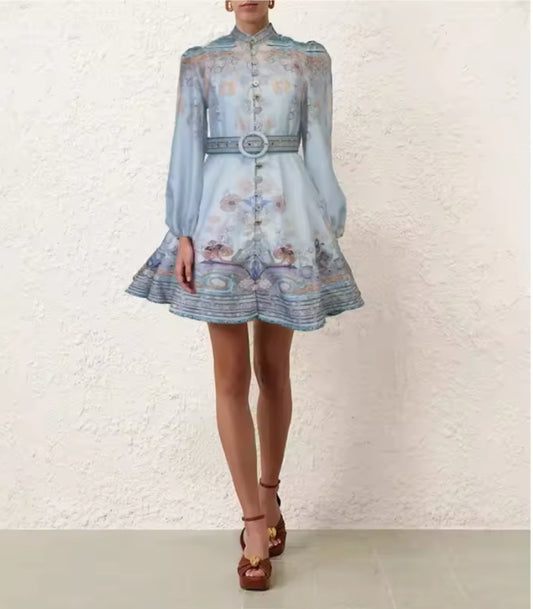 Model trägt ein pastellblaues Minikleid mit hohem Kragen, Knopfleiste, langen transparenten Ärmeln und floralem Muster, kombiniert mit passendem Gürtel