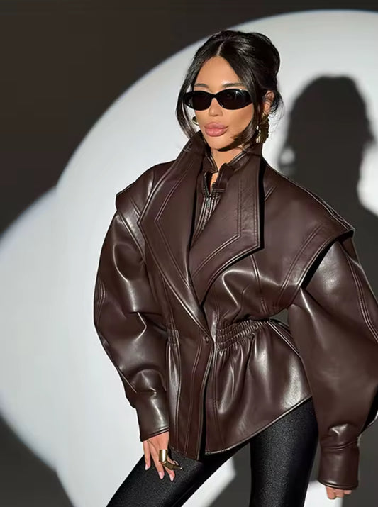 Braune Oversize-Lederjacke aus hochwertigem Kunstleder mit markanten Schultern, tiefem Reverskragen und Smok-Detail an der Taille – luxuriöser High-Fashion-Look.
