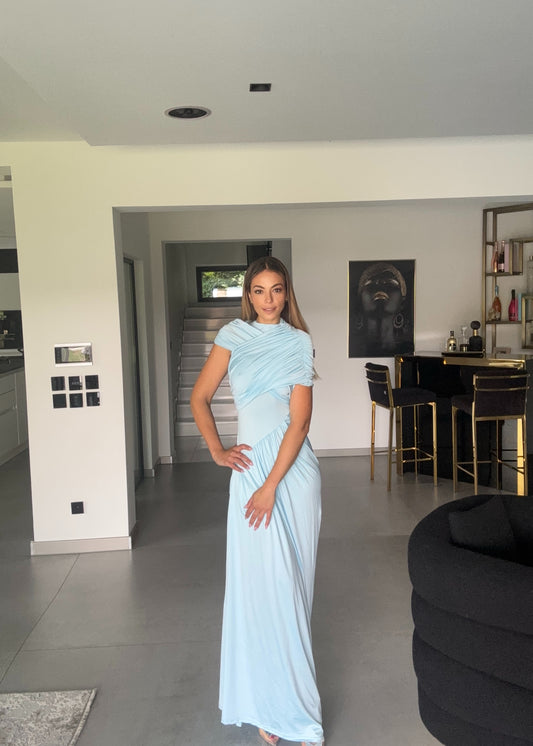 Frau trägt bodenlanges hellblaues Kleid mit drapiertem Oberteil und asymmetrischem Schnitt, steht in elegantem Interieur
